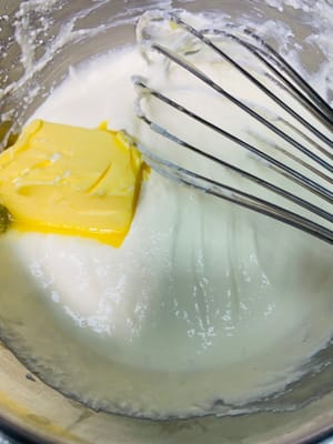 Die bei Zimmertemperatur geschmolzene Butter hinzufügen und weiterschlagen, bis eine glatte, partikelfreie Masse entsteht. Anschließend den Herd ausschalten, den Behälter aus dem heißen Wasser nehmen und etwas abkühlen lassen.