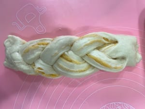 240g椰蓉辫子包:
把面团擀平,铺上椰蓉酱,折好再轻轻压扁。
用刀切成三条(顶端别切断),有椰蓉的一面朝上,像编辫子一样编起来,收口捏紧。
把两端向里卷成一个圆,放进240g的一次性面包盒里发酵。