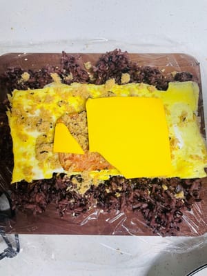 將準備好的食材中最大片的放在底層,然後依次往上堆疊,最後放上芝士片、擠上沙律醬並塗抹均勻。依照圖示箭頭方向,兩手提起保鮮膜往中間收緊,再稍用力拉緊,即可捲成一個長條狀的飯糰。