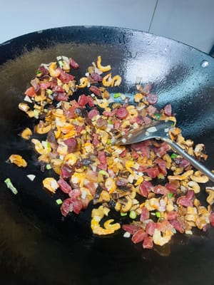 Öl in einer Pfanne bei niedriger Hitze erhitzen. Zwiebeln und die weißen Teile der Frühlingszwiebeln hinzufügen und anbraten, bis sie duften. Dann die getrockneten Garnelen dazugeben und kurz anbraten, bis sie duften. Anschließend das gepökelte Schweinefleisch und die Wurst dazugeben und anbraten, bis das Fett ausgelassen ist. Nicht zu stark erhitzen, da das Fleisch sonst trocken und zäh wird. Zum Schluss die Shiitake-Pilze dazugeben und einige Minuten mitbraten.