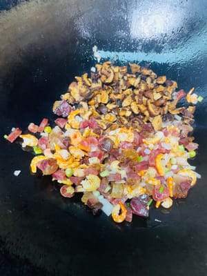 Öl in einer Pfanne bei niedriger Hitze erhitzen. Zwiebeln und die weißen Teile der Frühlingszwiebeln hinzufügen und anbraten, bis sie duften. Dann die getrockneten Garnelen dazugeben und kurz anbraten, bis sie duften. Anschließend das gepökelte Schweinefleisch und die Wurst dazugeben und anbraten, bis das Fett ausgelassen ist. Nicht zu stark erhitzen, da das Fleisch sonst trocken und zäh wird. Zum Schluss die Shiitake-Pilze dazugeben und einige Minuten mitbraten.