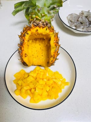 Ananas vorbereiten: Zuerst eine Seite der Ananas so dünn schneiden, dass sie gut auf dem Teller liegt. Von der anderen Seite etwa 1/3 abschneiden. Anschließend mit einem Messer mehrere horizontale und vertikale Schnitte in die Ananas machen (wie ein Gitter). So lässt sich das Fruchtfleisch leicht herauslöffeln. Das Fruchtfleisch würfeln, die Schale bleibt als „natürlicher Behälter“ für Reis erhalten – ideal für spätere Verwendung in gebratenem Reis.